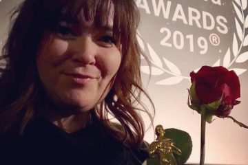 Heli Järvenpää hymyilee palkintopysti ja ruusu kädessä. Taustalla valkokangas, jossa heijastettuna "eOppiva Awards 2019" -tunnus.